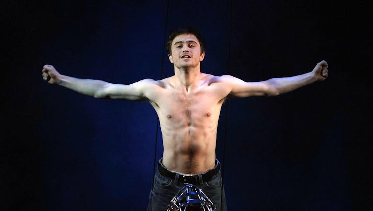 A young man, shirtless, on stage against a black background.