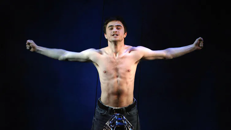 A young man, shirtless, on stage against a black background.