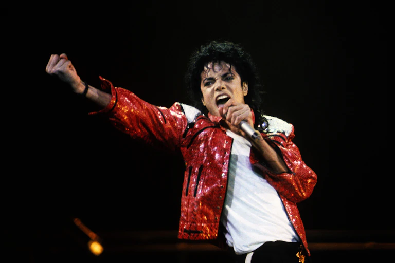 Michael Jackson sings with a fist raised. He wears a sparkling red jacket over black pants and a white T-shirt.