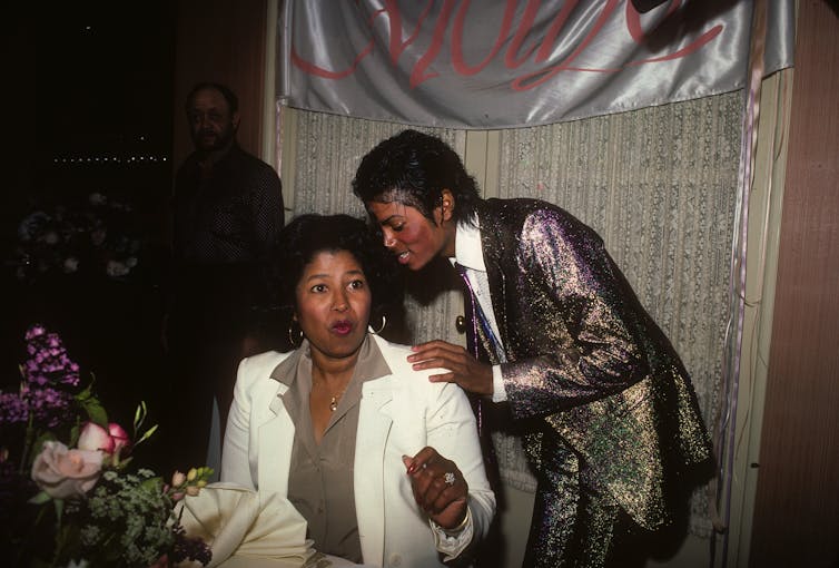 Katherine Jackson, wearing a white jacket over an olive-green shirt, is seated at a table covered with flowers; her son Michael Jackson stands behind her, wearing a sparkly suit.