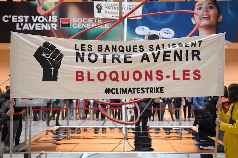 Des manifestants tiennent une banderole sur laquelle on peut lire « Les banques salissent notre avenir. Bloquons-les » lors d'une action de protestation à l'entrée de la banque Société Générale dans le quartier d'affaires de La Défense, à l'ouest de Paris, le 15 mars 2019 en marge de la manifestation mondiale des jeunes pour le climat.