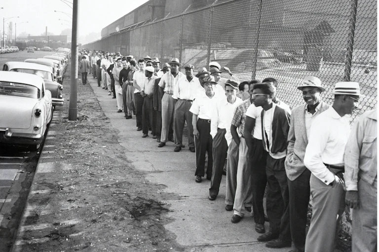Steelworkers in casual clothes lineup outside the South Works mill. The line is a mix of a Black, white and Hispanic workers.