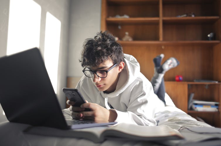 A teenage boy lies on a bed with a laptop and book, while testing on a phone.
