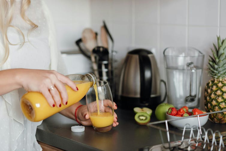 A woman pours orange juice into a glass next to a bowl of strawberries and kiwifruit.