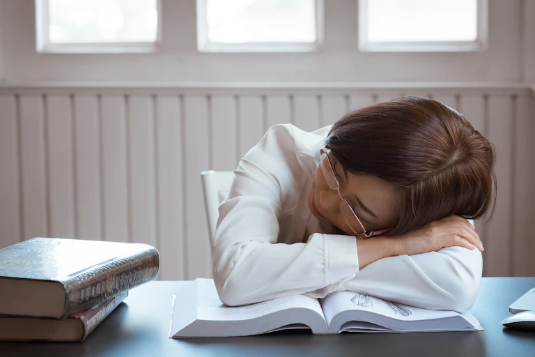 A girl in glasses rests her head on an open textbook.