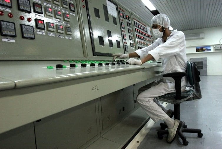 Um técnico de terno branco e máscara está sentado em um enorme console com vários botões e mostradores.