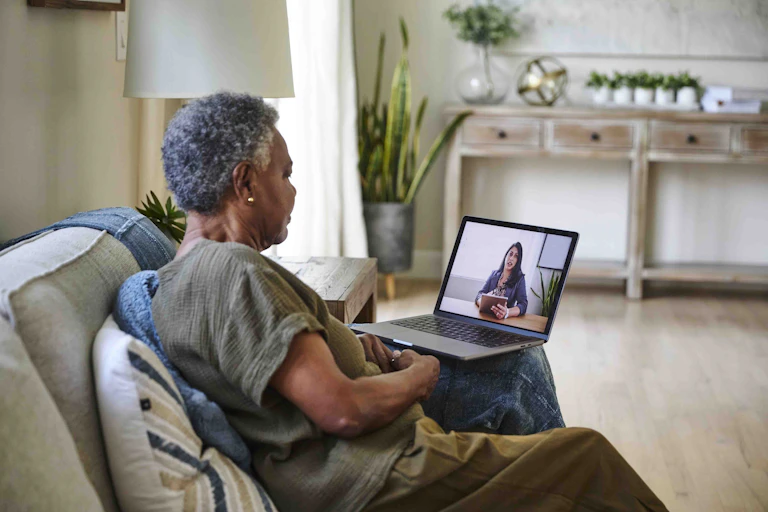 A woman with gray hair sits on a couch during a videocall with a brunette woman holding a clipboard.