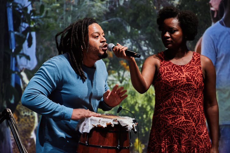A woman holds a microphone to a man playing a drum