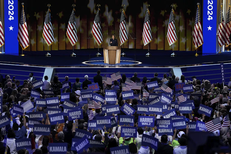A woman stands on a stage with rows of American flags behind her and crowds of people in front of her waving Kamala signs.