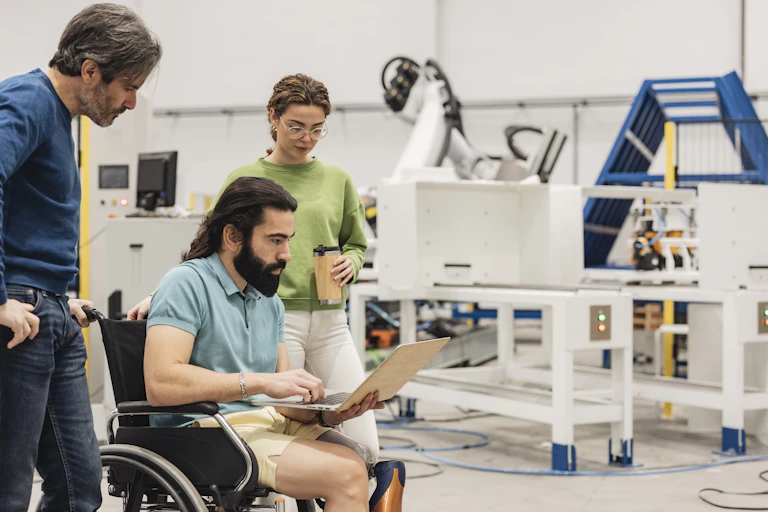Engineer sitting in wheelchair discussing with colleagues on laptop in factory.