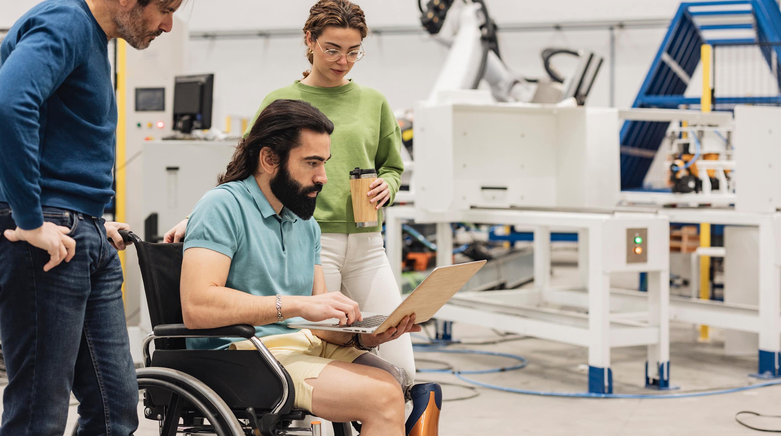 Engineer sitting in wheelchair discussing with colleagues on laptop in factory.