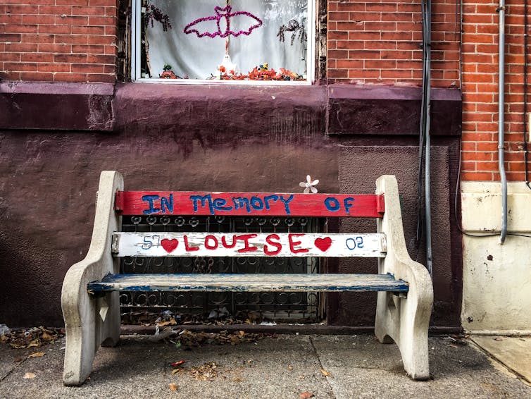 A park bench with the words 'In Memory of Louise' written on it sits in front of a brick row home