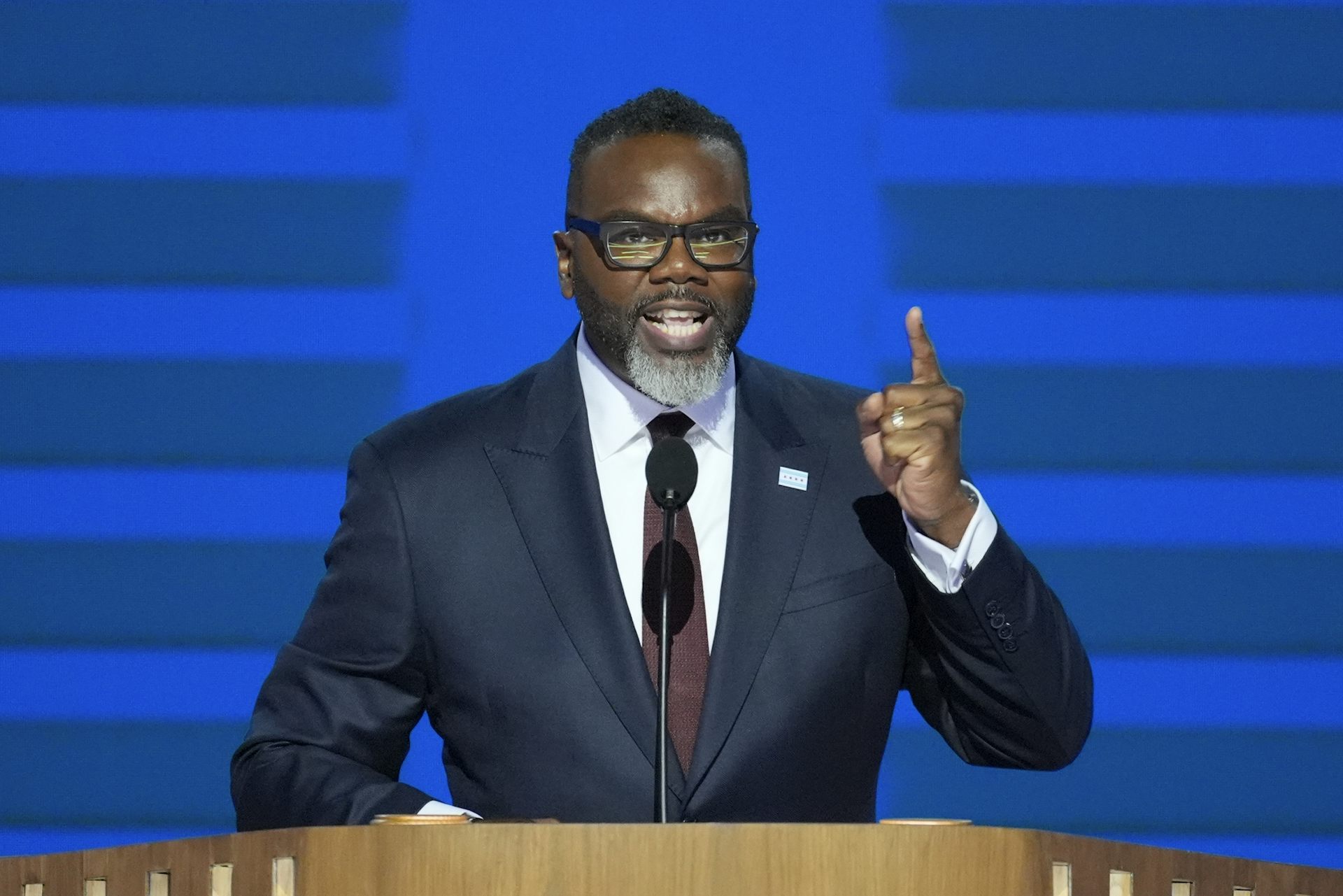A Black man in a dark suit gestures while speaking from behind a lectern.