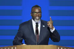 A Black man in a dark suit gestures while speaking from behind a lectern.