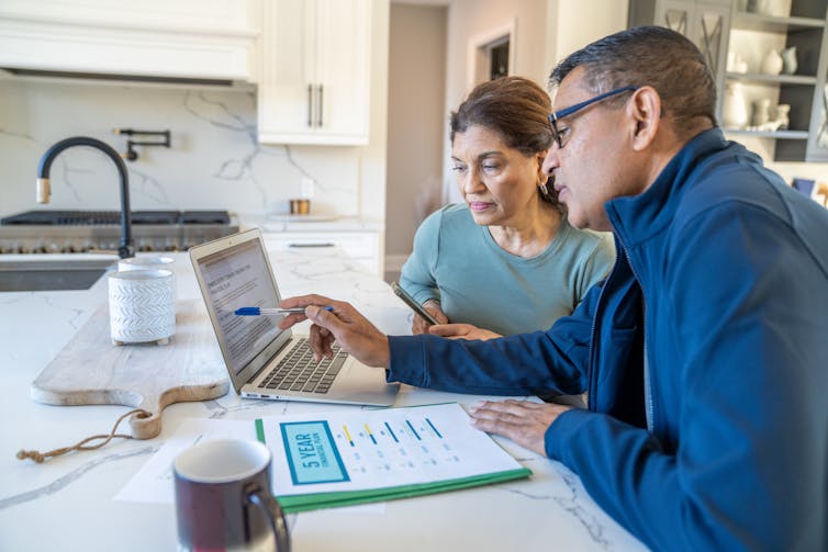 A couple sit at their kitchen table with a laptop open and their bills spread out in front of them.