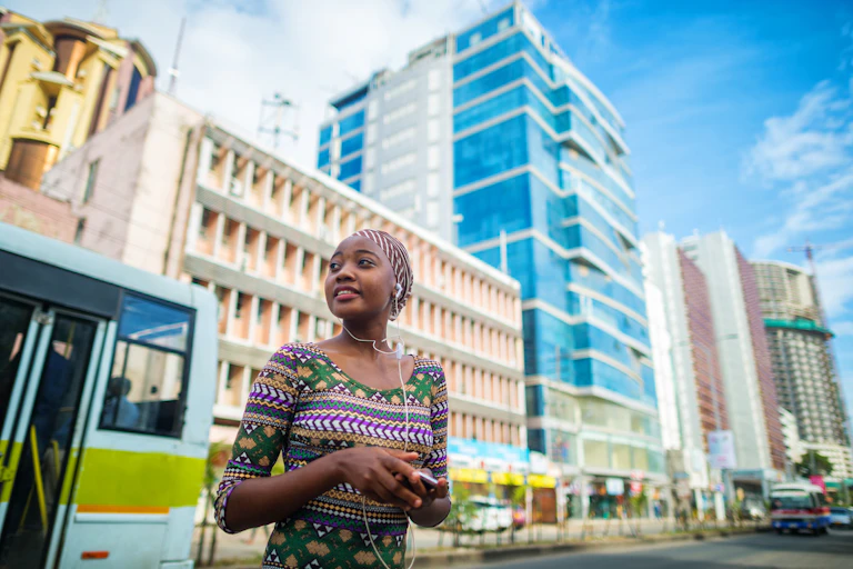 Uma mulher jovem e de aparência confiante está em uma rua de aparência próspera em Dar es Salaam. Ela usa fones de ouvido brancos e um top multicolorido; atrás dela há um prédio de vidro reluzente e um ônibus.