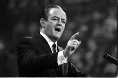 A black and white photo shows a man with thinning hair in a dark suit pointing as he speaks into a microphone.