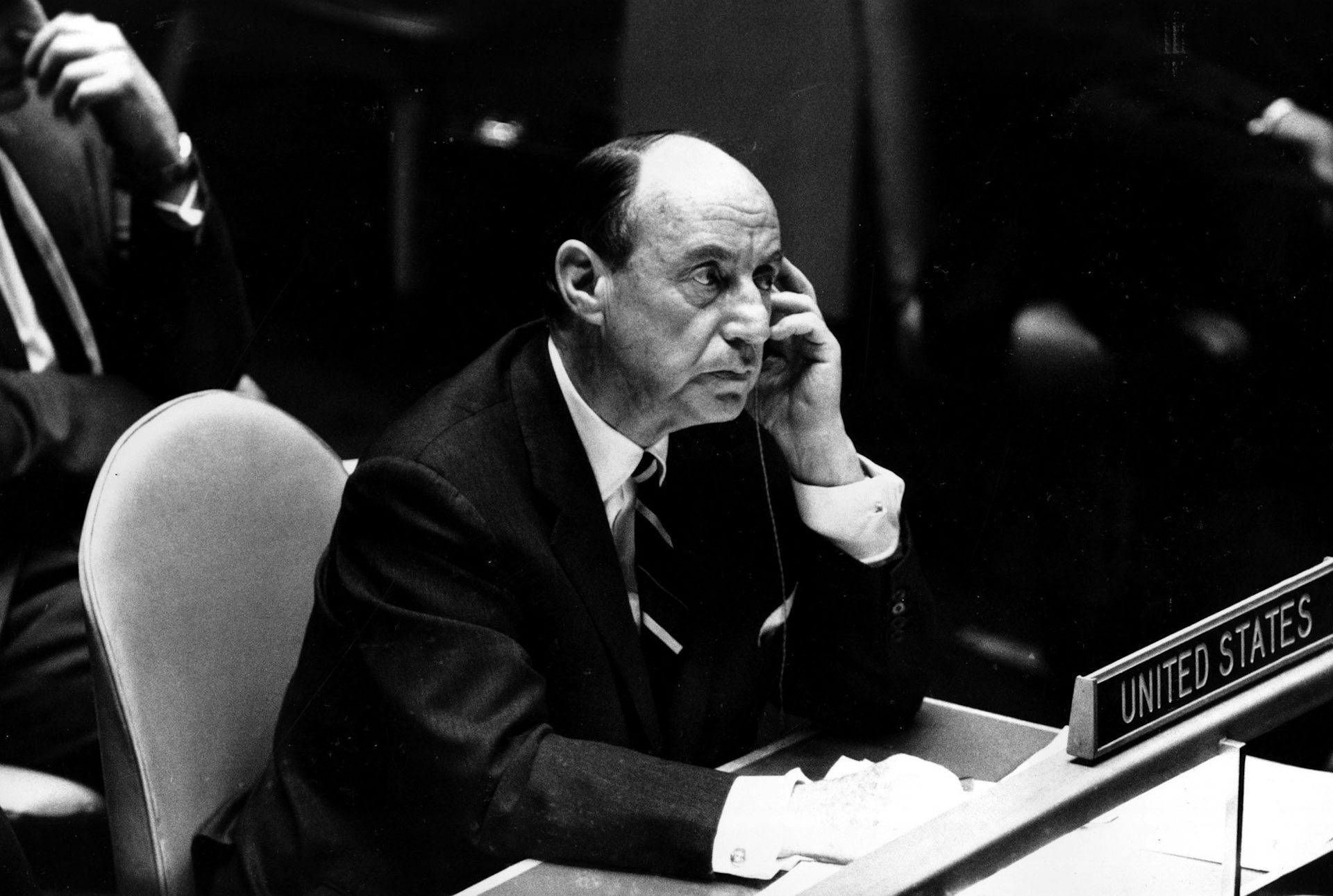 A black and white photo shows bald man in a dark suit holding a telephone receiver in his left hand.