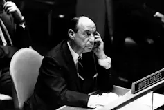 A black and white photo shows bald man in a dark suit holding a telephone receiver in his left hand.