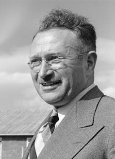 Black and white headshot of a middle-aged man who has a mustache and is wearing glasses.