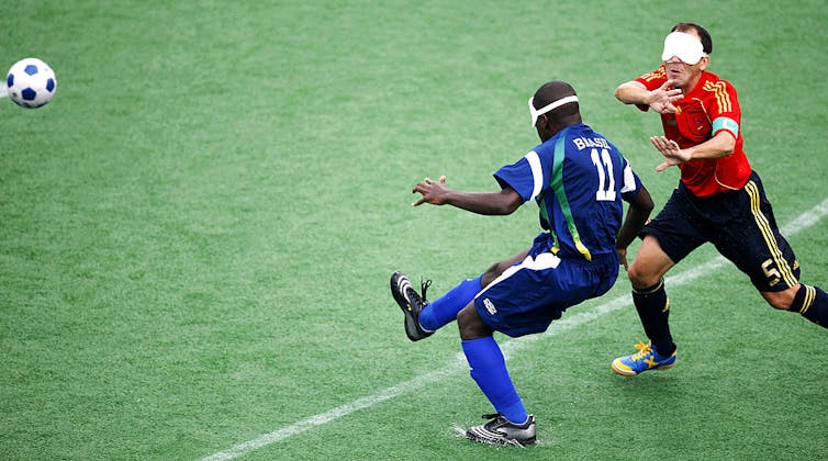 Young man wearing blue jersey and white eye mask kicks ball as player wearing red jersey runs toward him.