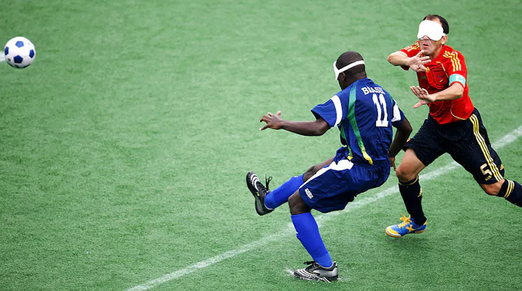 Young man wearing blue jersey and white eye mask kicks ball as player wearing red jersey runs toward him.