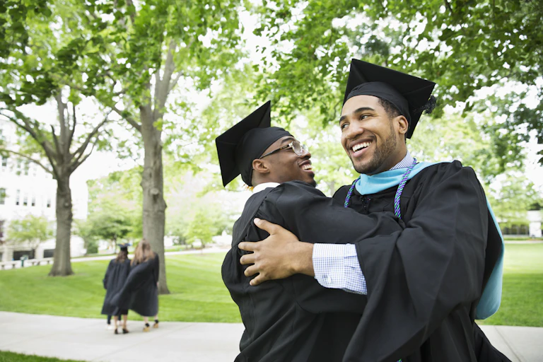 Wearing caps and gowns, two Black male college graduates embrace each other.