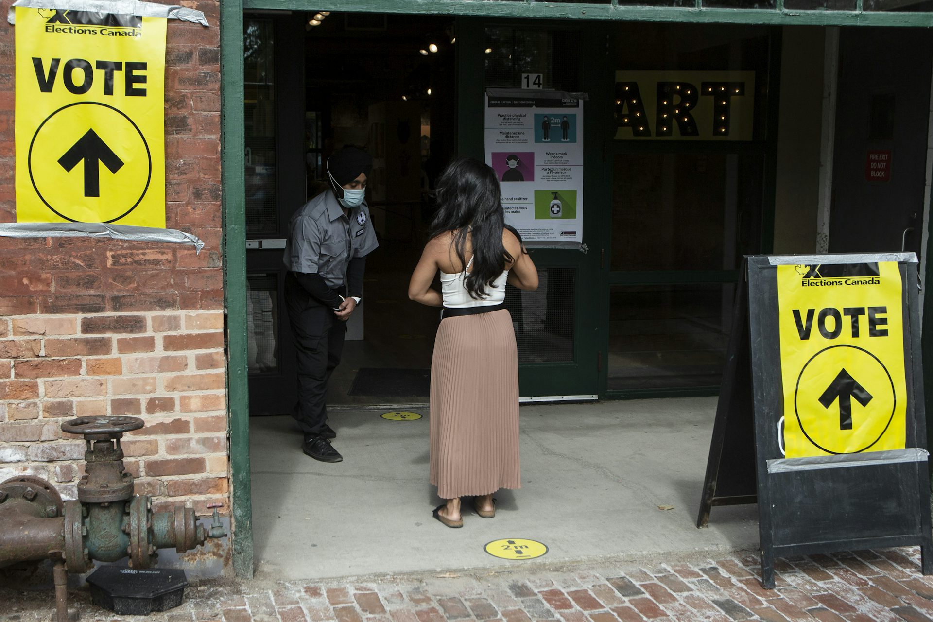 A woman with long dark hair and wearing a white tank top and long beige skirt is photographed from behind entering a polling station.