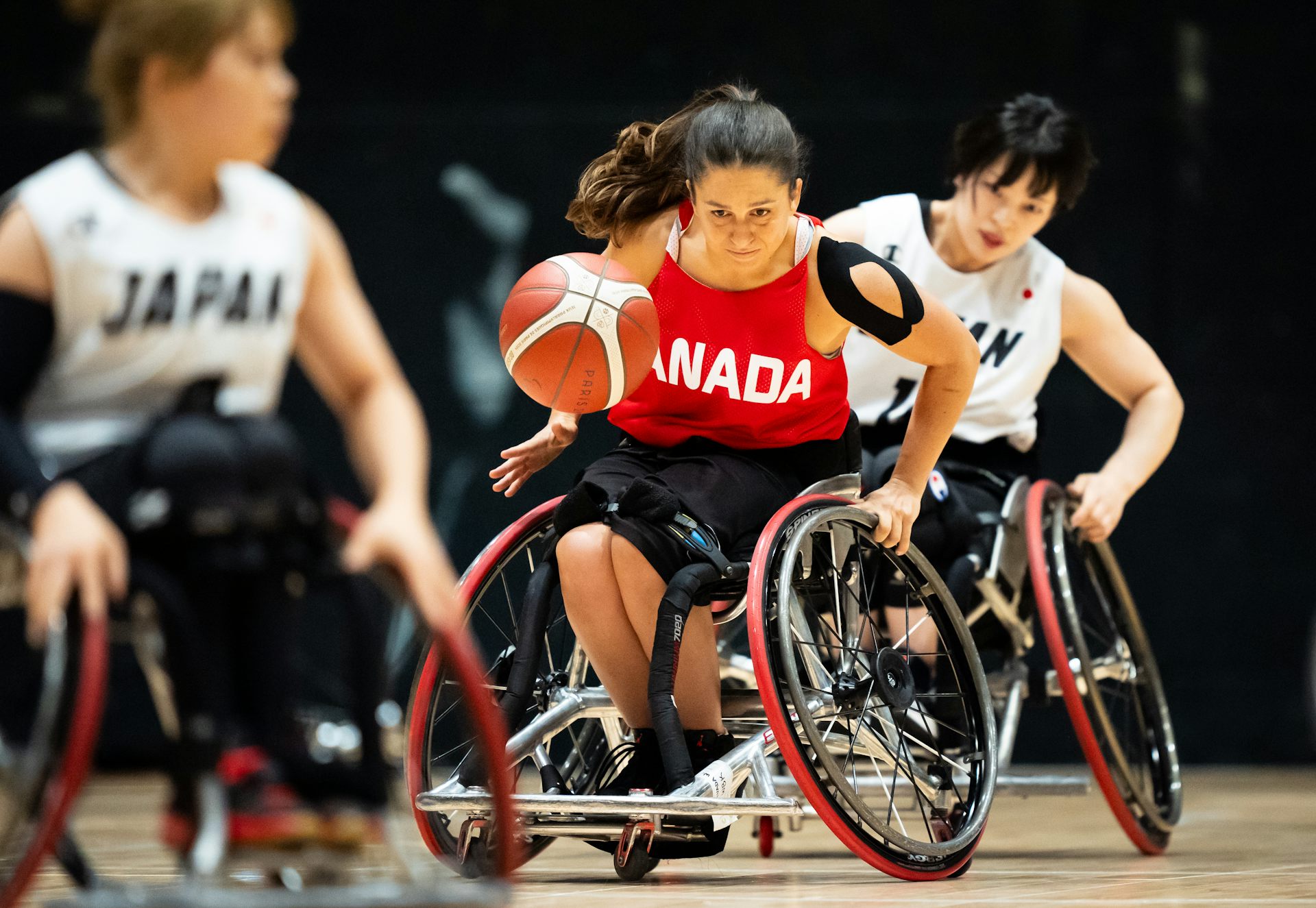 Three women, one in a red Canada jersey and two in white Japan jerseys, playing wheelchair basketball.