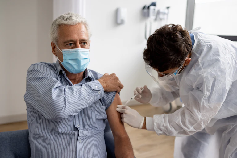 A male doctor in a hospital gives an adult man with white hair a shot in his left arm.