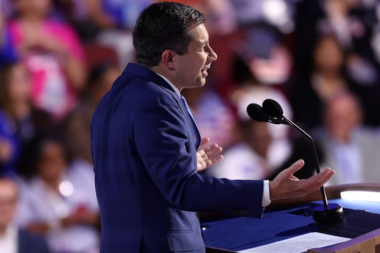 A man in a blue suit jacket, standing at a microphone and gesturing with his hands.