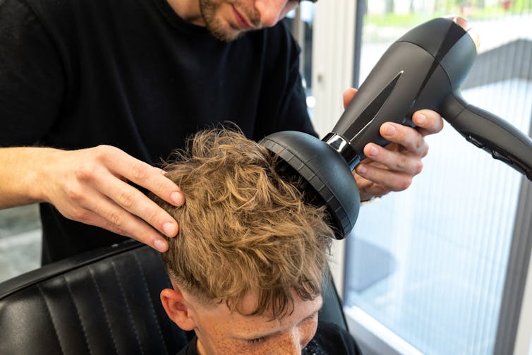 A man uses a diffuser attachment on a hair dryer to give a male client curly hair.
