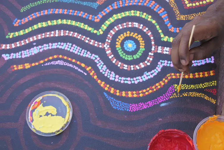 A hand paints an intricate Aboriginal dot painting in many colours