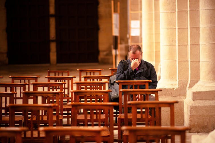 A lonely man sitting in a row of chairs in a church with his head in his hands