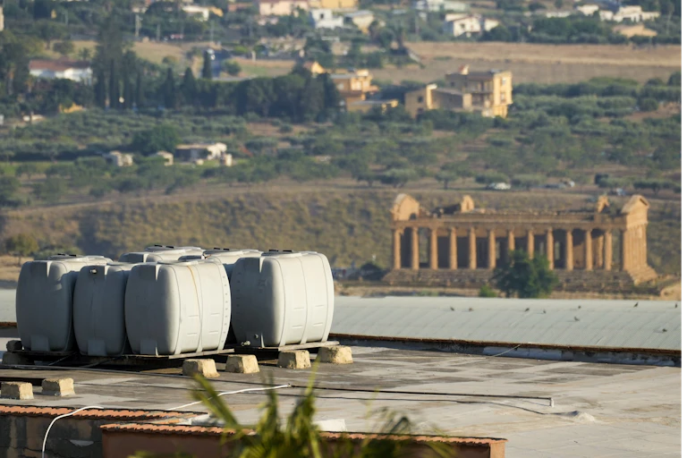 Water containers are seen on a roof with ruins in the background.
