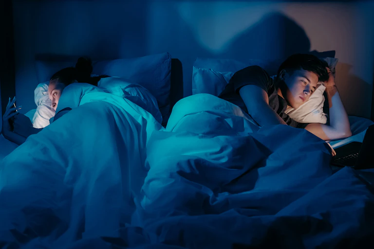 Man working using laptop in bed at night while partner is turned away using phone.