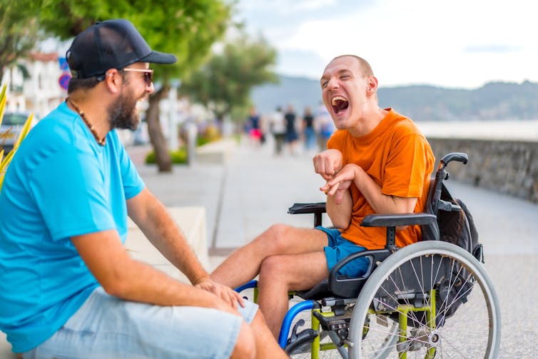 A person in a wheelchair laughs with a friend on a boardwalk