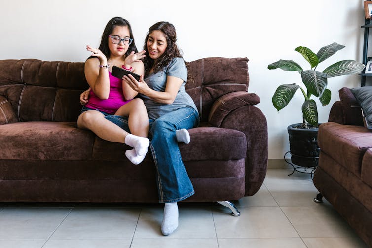 Two women, one with disability, sit on a couch looking at a phone