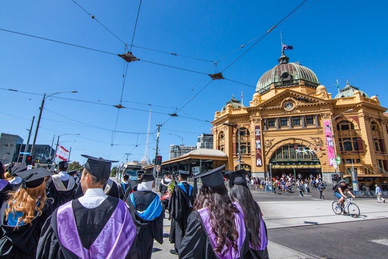 Graduates in caps and gowns walk across the road. Tram lines are overhead.