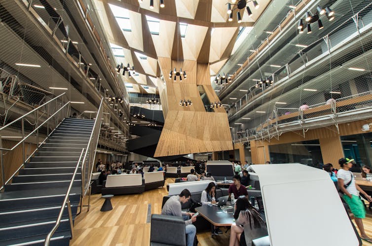 Young people work on computers in small clusters of desks in a large lobby. There is a high ceiling with lights and a wide staircase.