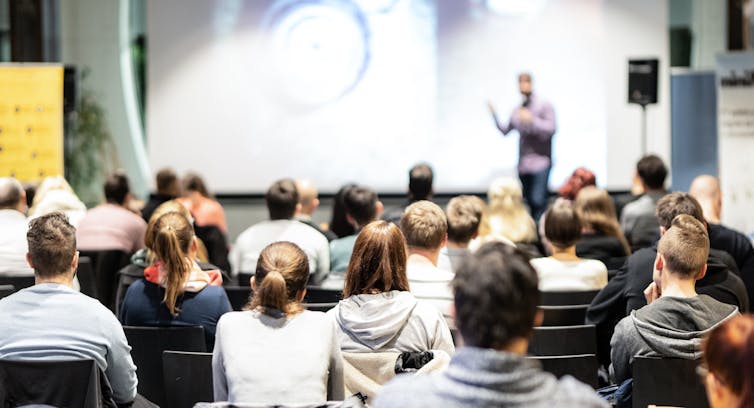 Rows of people in seats listen to a speaker, who is standing in front of a whiteboard, next to a speaker.
