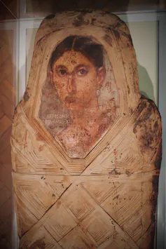 A human body shaped object wrapped in linen, with a detailed dark haired woman's portrait painted on it.