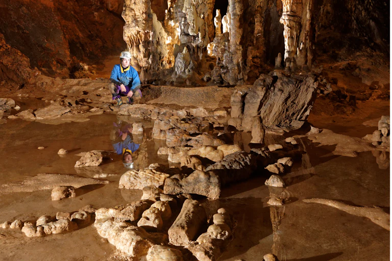 Dans une grotte, une jeune femme accroupie observe des roches brisées et installées sur le sol.