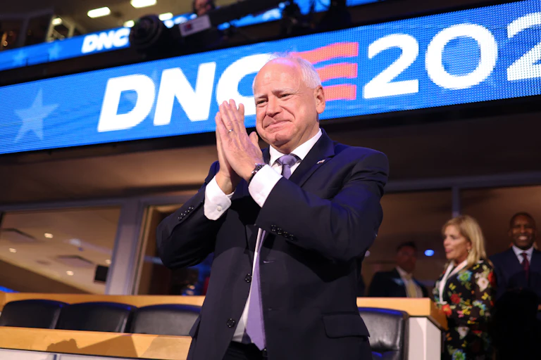 A man in a suit smiles as he holds his hands in front of his face.