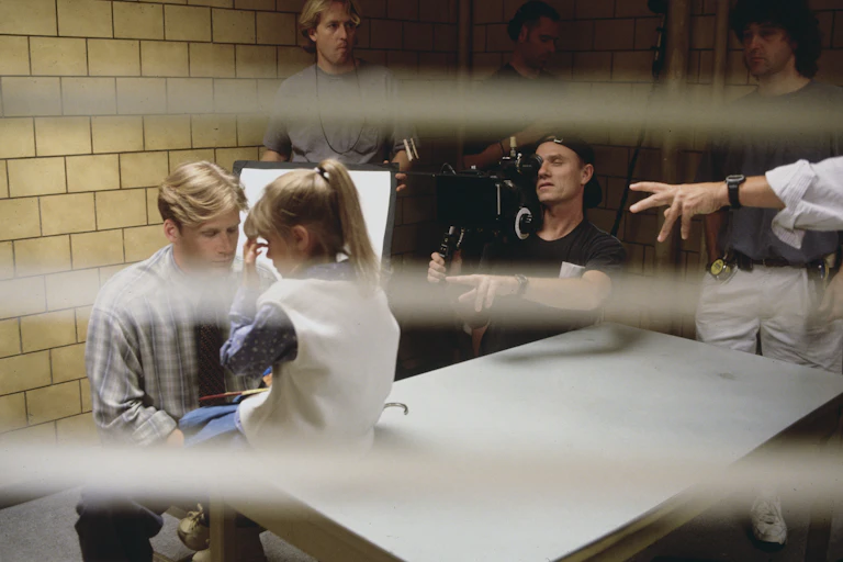 Young man talks to young girl sitting on table while a man films them with a big, black camera.