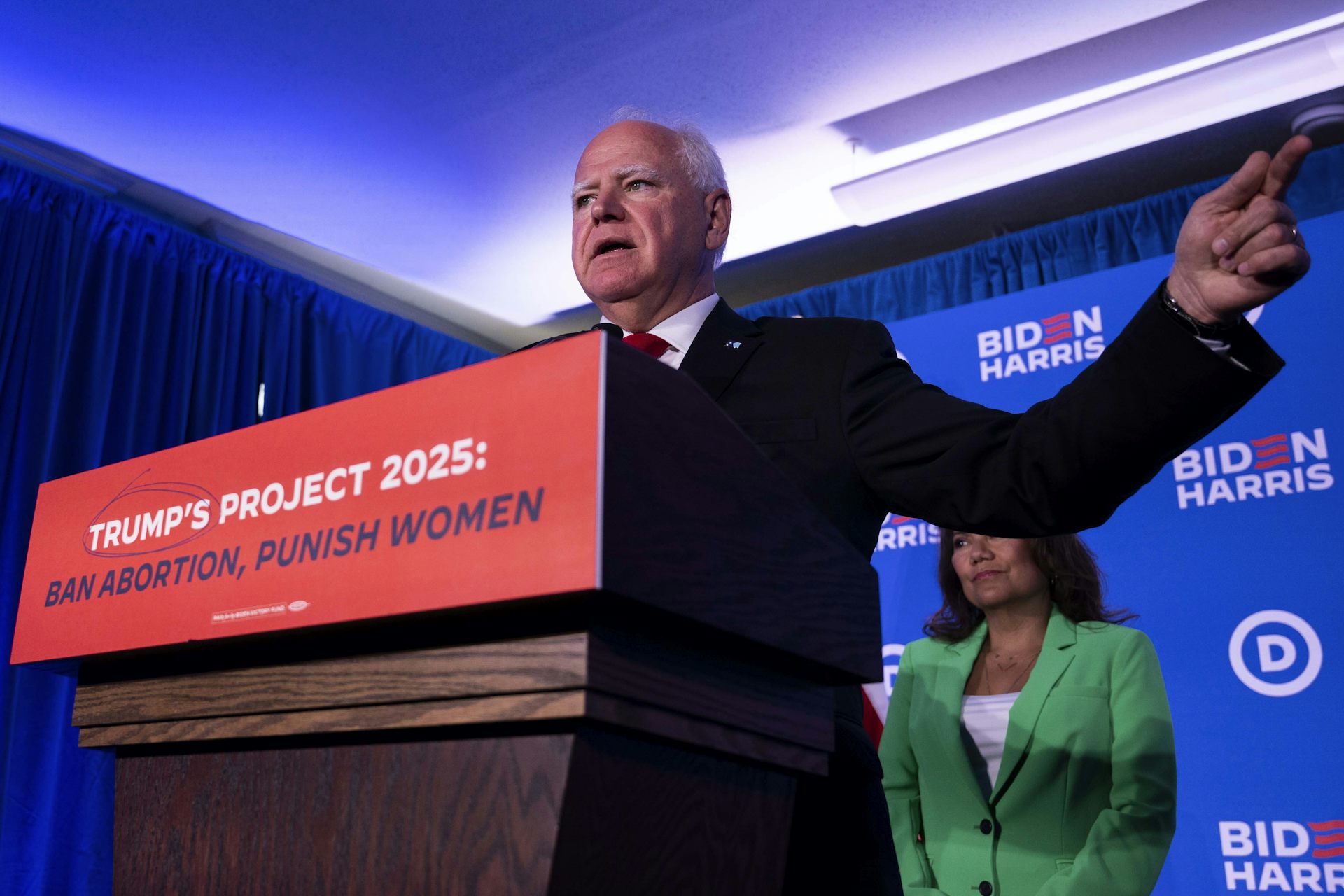 A man stands at a lectern gesturing. A banner at the front of the lectern says: Trump's Project 2025: Ban abortion, punish women.