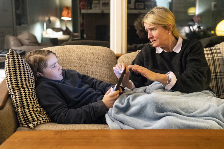 concerned woman taking a tablet from a grumpy child lying on the couch
