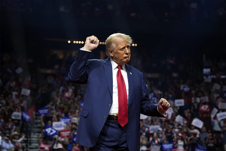 A heavily tanned man in a blue suit and red tie with grey-blond hair dances awkwardly in front of a crowd.