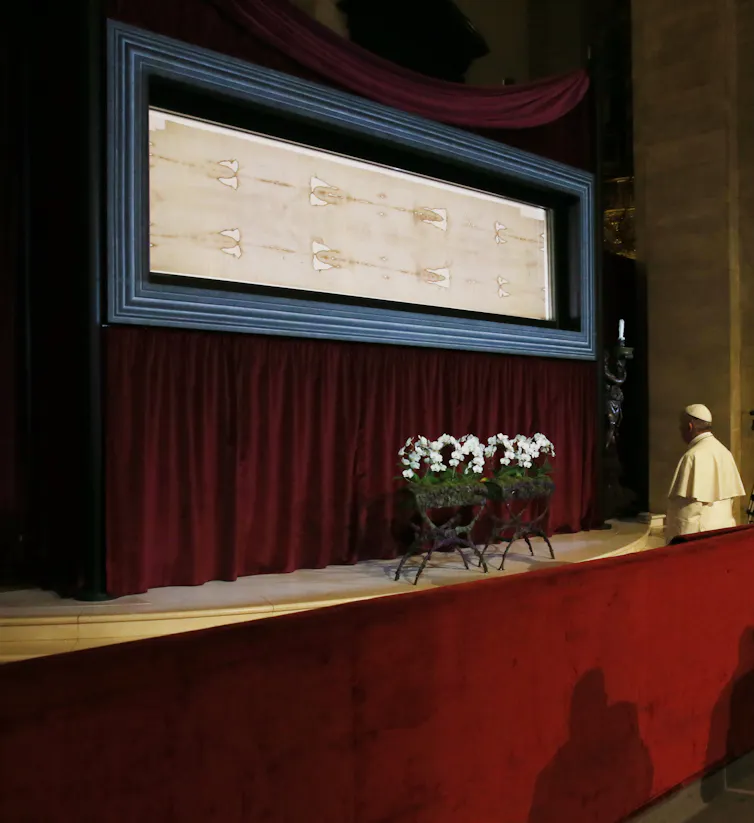 A man in a white robe looks intently at a framed panel containing a yellowed print with long lines, slightly tattered at the edges. A figure is faintly visible in the background.