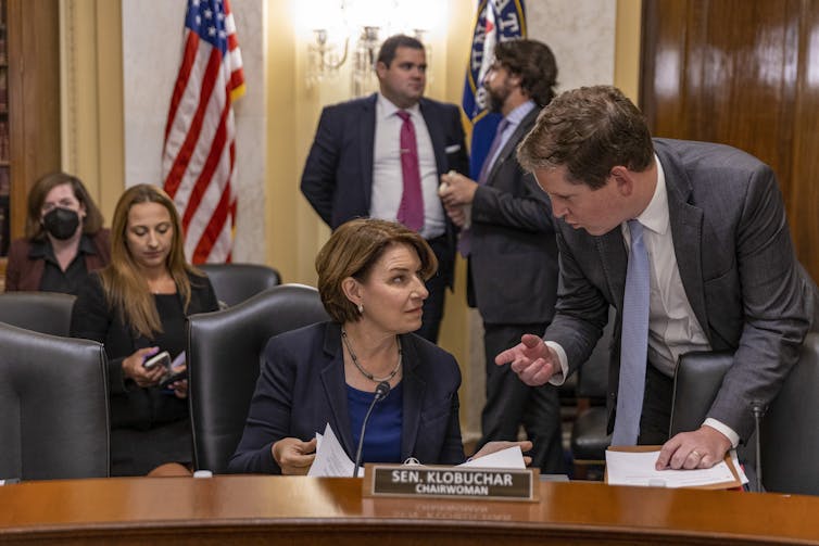 A woman in a jacket and blue dress talks to someone as she sits at a desk, with flags on poles behind her.